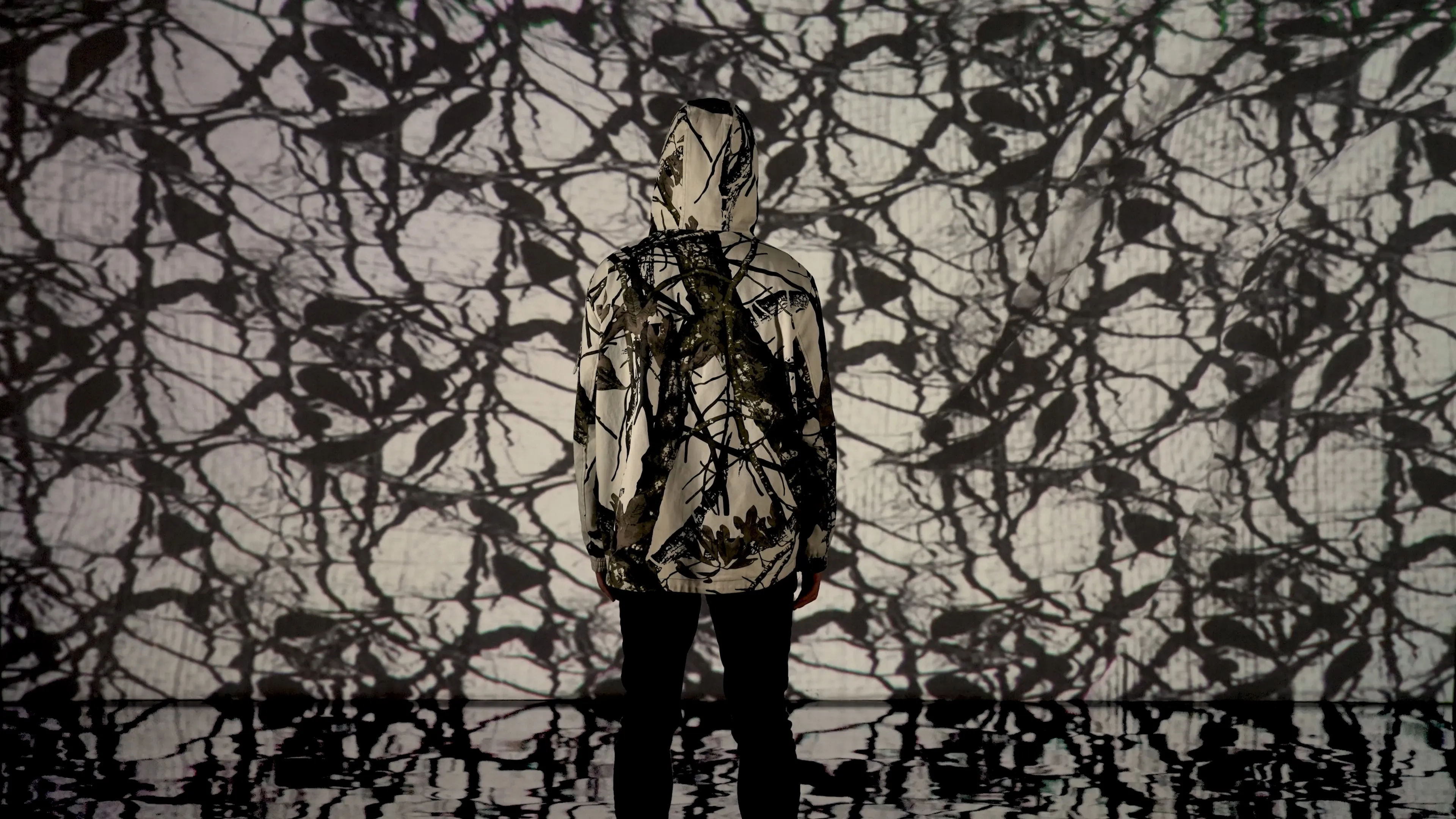 A person in a hoodie featuring a tree branch pattern stands in front of a projection screen displaying a similar pattern, created with generative AI.
