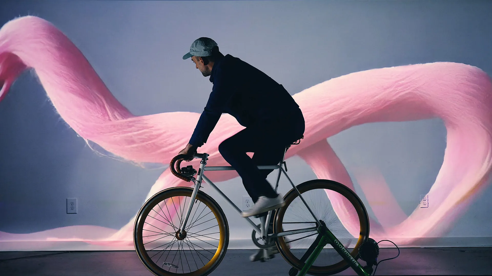 Interactive art installation with a stationary bike connected to a digital screen displaying ai generated abstract graphics that change in response to the cyclist's movements. The graphics look like a long string of pink cotton candy on a blue background.