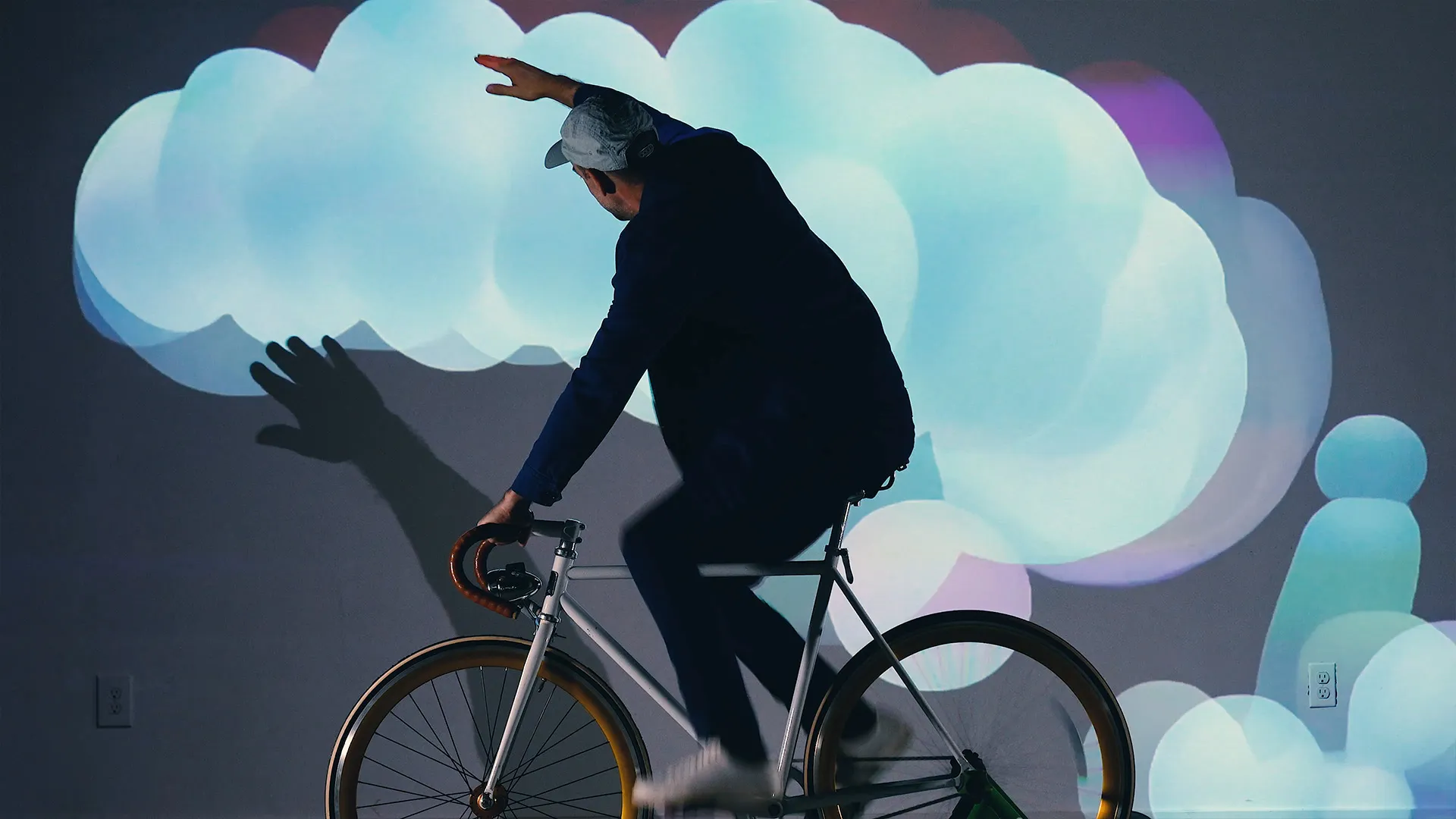 The rider on the stationary bike in fornt of a large projection lifts his hands in the air. A kinect sensor is mounted above the bike to track the rider's movements, triggering additional animations based on the position of the rider's hands.