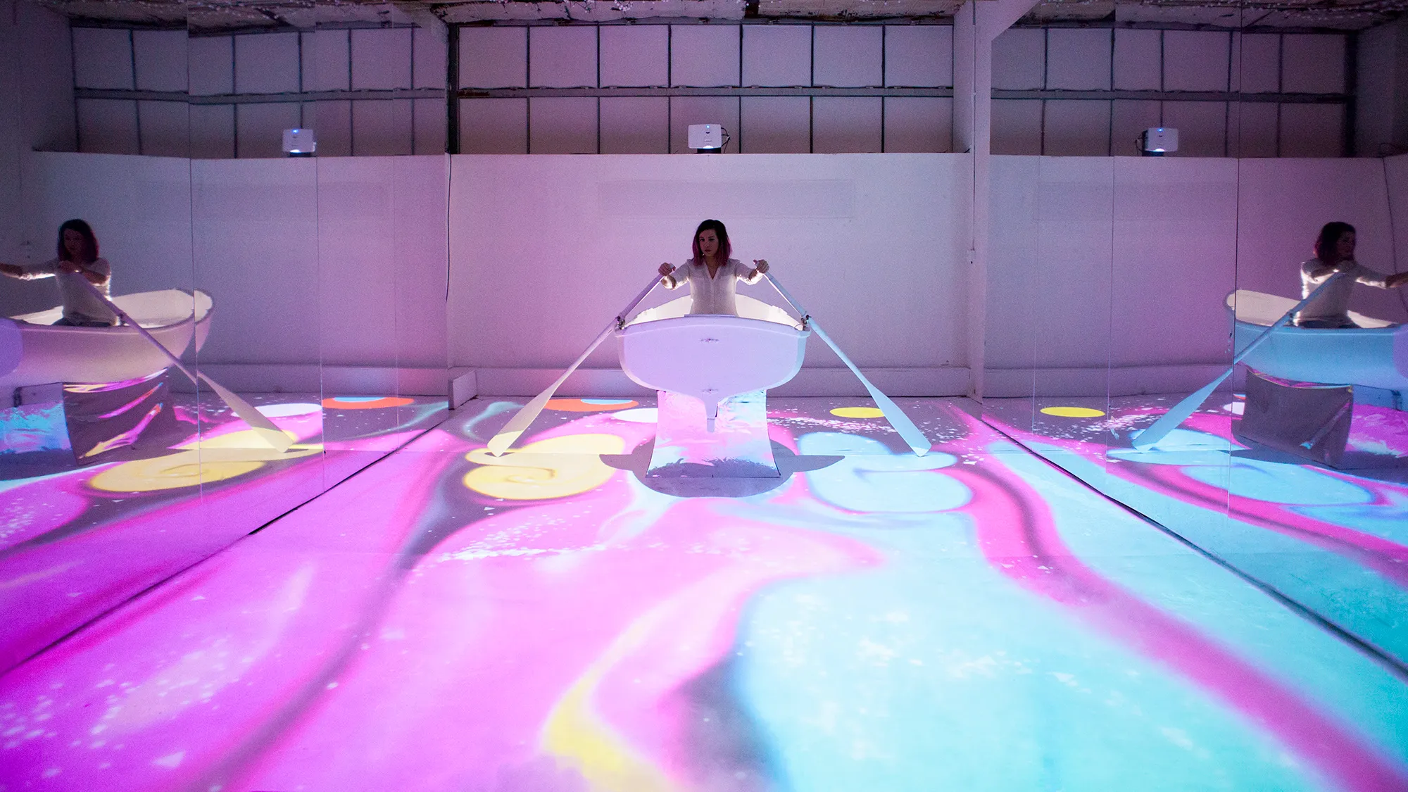 A girld in a boat, rowing through magical, floor projected graphics that are influenced by fluid dynamics.