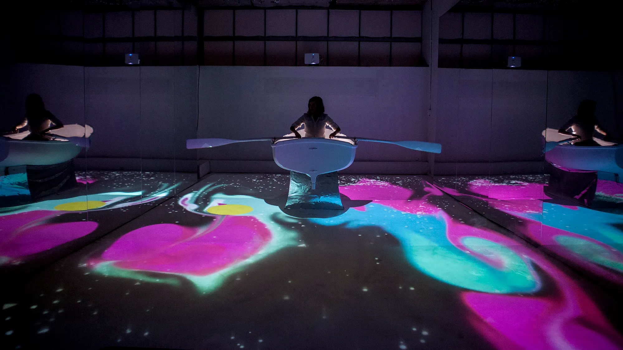 A girld in a boat, rowing through magical, floor projected graphics that are influenced by fluid dynamics.