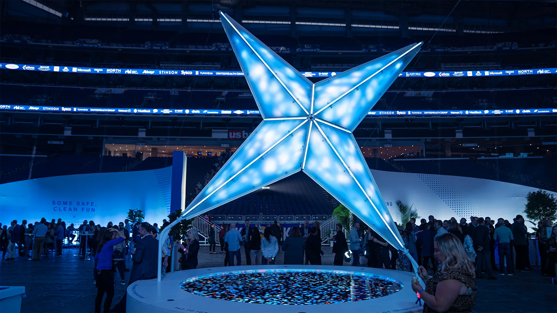 The LED sculpture radiates light from the center, its base LED screen displaying dynamic particle visuals. Around it, event attendees gather in a semicircle, captivated by the display.