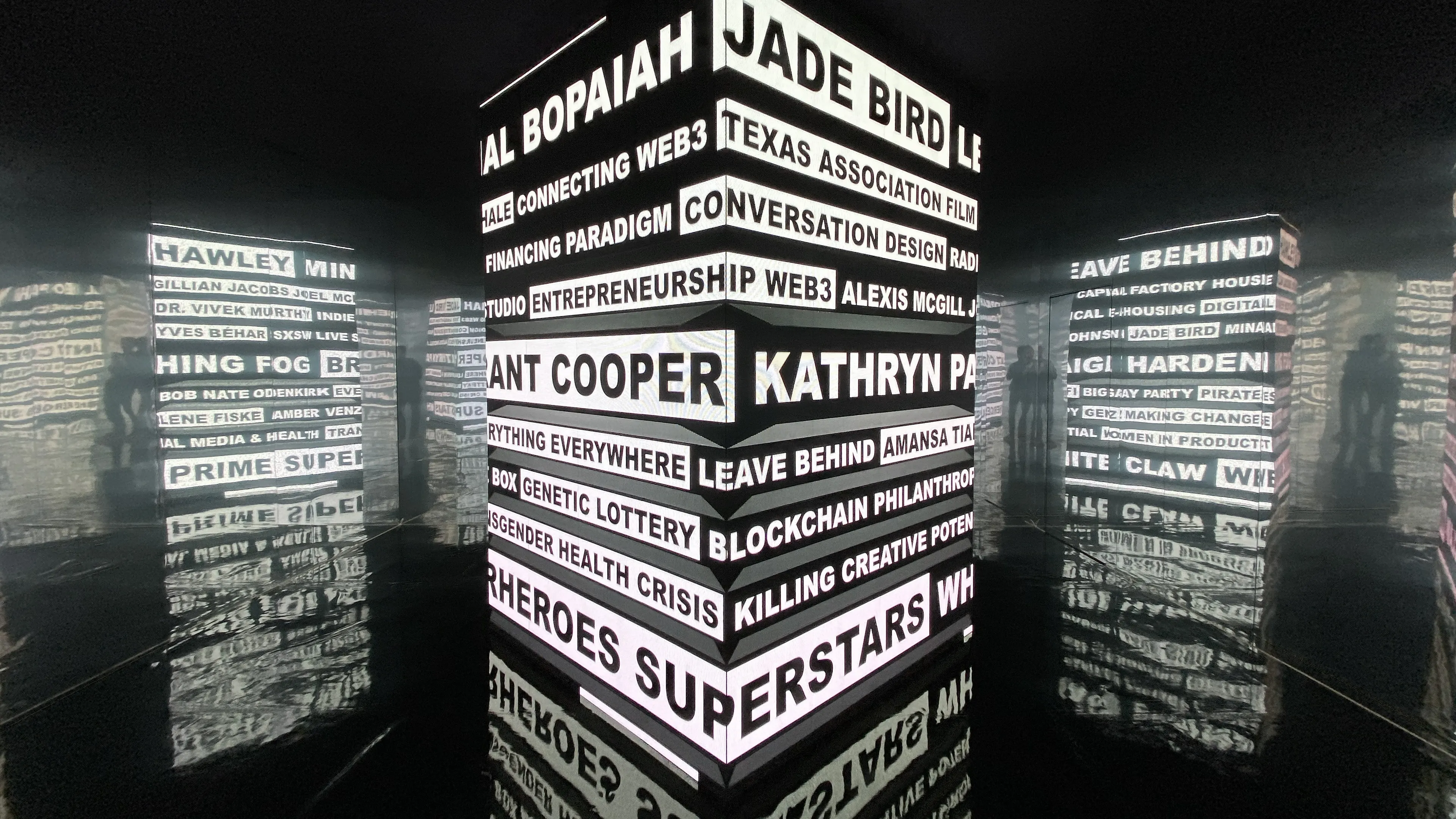 An interactive art installation showcases an LED cube at the center of a room encased in mirrored walls. The cube cycles through black and white typography, prominently listing the names of bands featured at SXSW 2022.