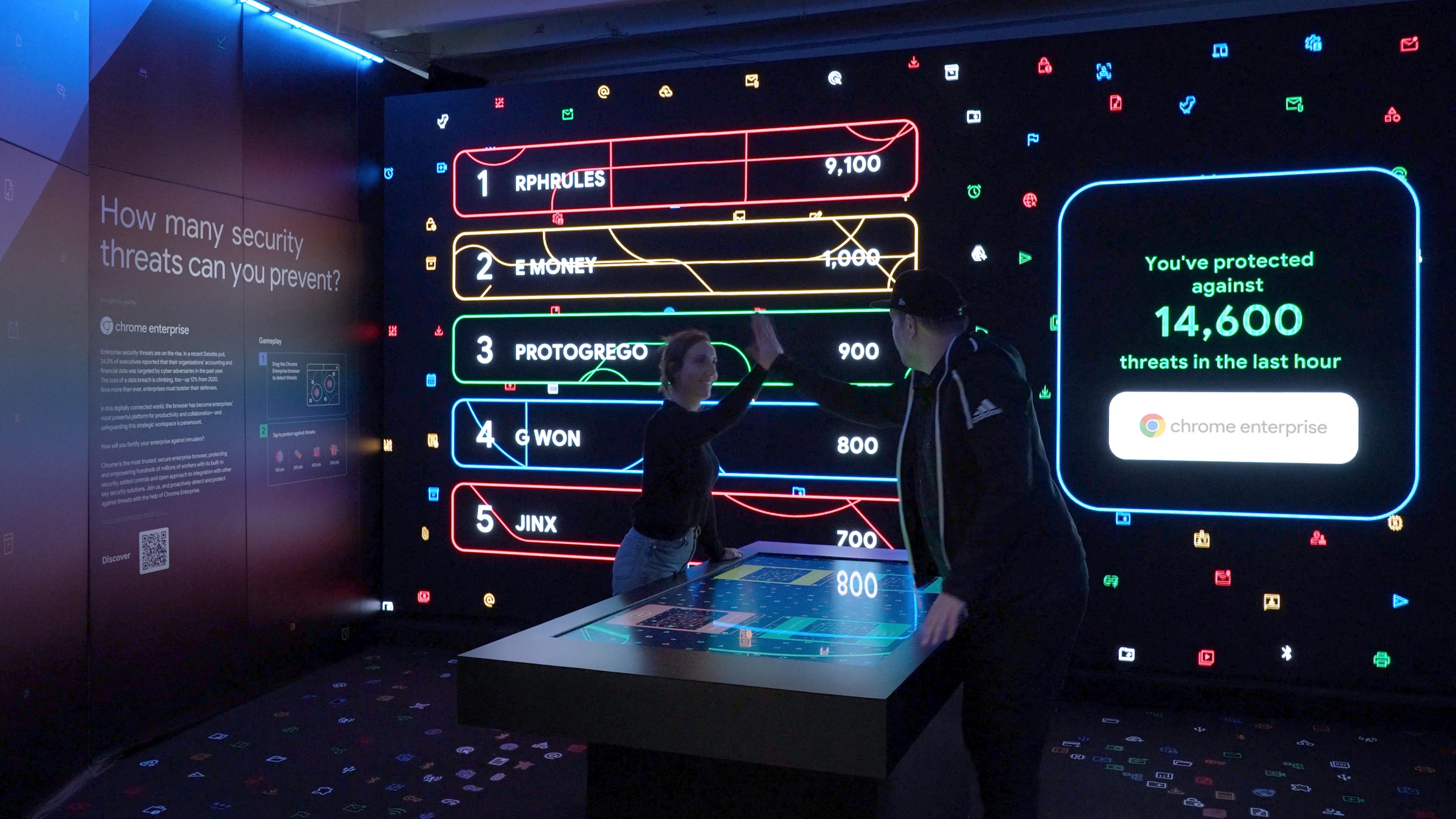 Two players high fiving each other across the touchscreen table after finishing a successful game of Data Defense. The Big LED Wall behind them shows Data Defense's leaderboard.