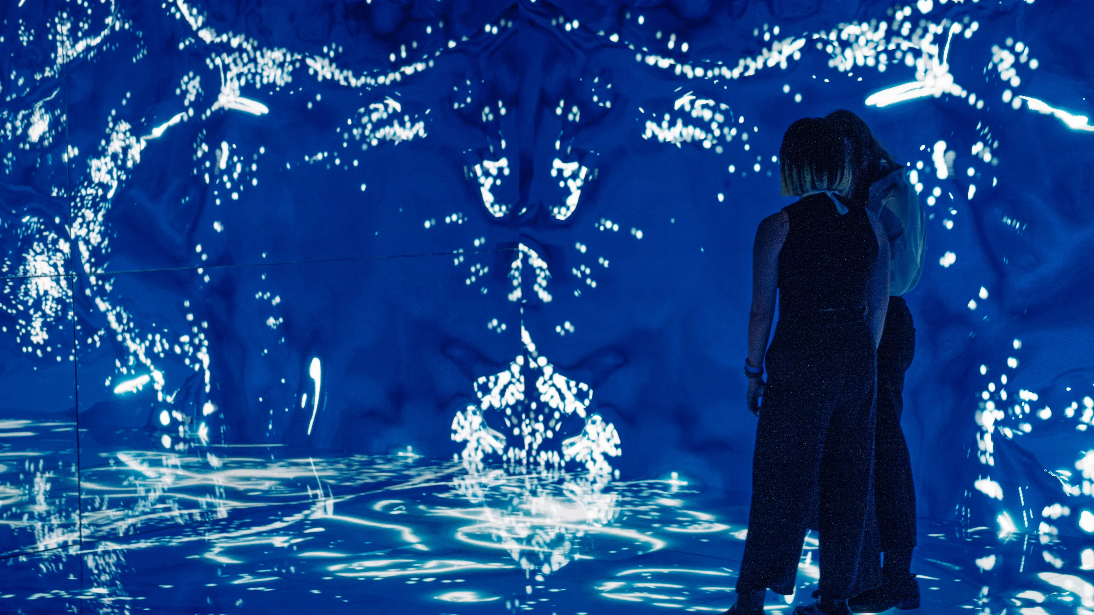 Two people standing in an interactive installation, blue water graphics surround them on the walls and floor, mirrored walls reflect the graphics