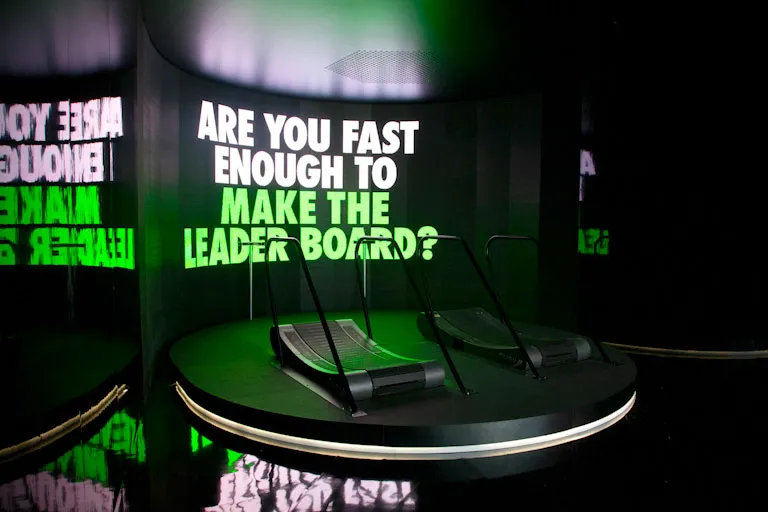 Treadmill game setup: two empty, self powered treadmills on a stage in front of a large LED screen that invites players by displayig the questions: Are you fast enough to make the leaderboard?