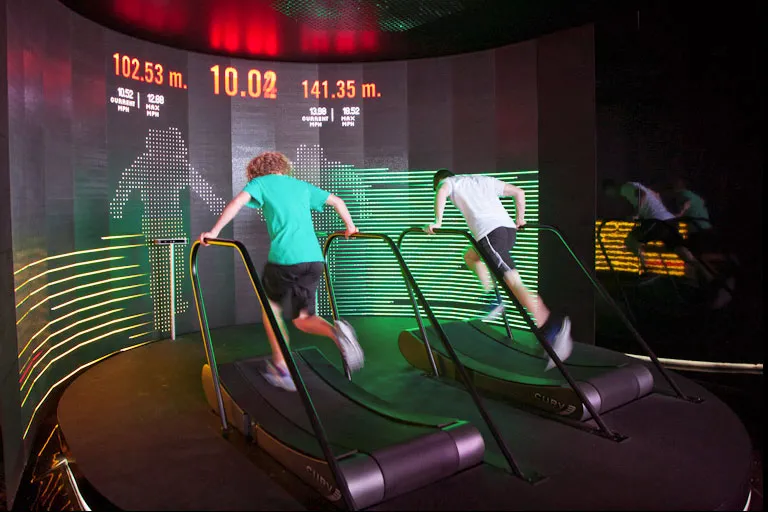 Treadmill game gameplay: two players running on treadmills in front of a large LED screen that displays their silhouettes, distance ran and a countdown.